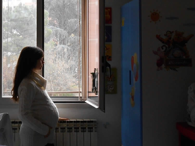 Images shows a pregnant woman looking out of a window.