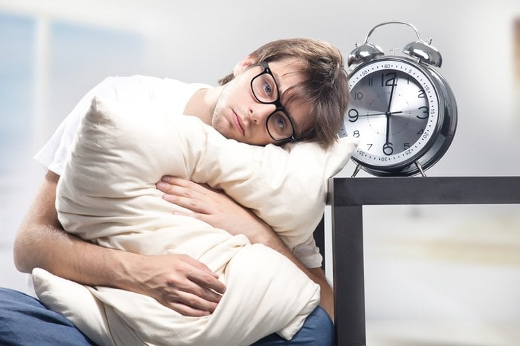This image shows a tired looking man holding a pillow and sitting next to an alarm clock.