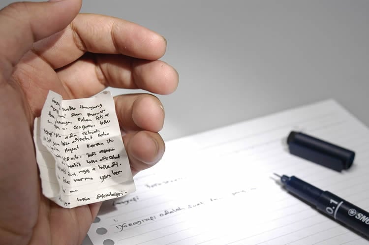 This image shows a hand holding cheat notes and an exam paper.