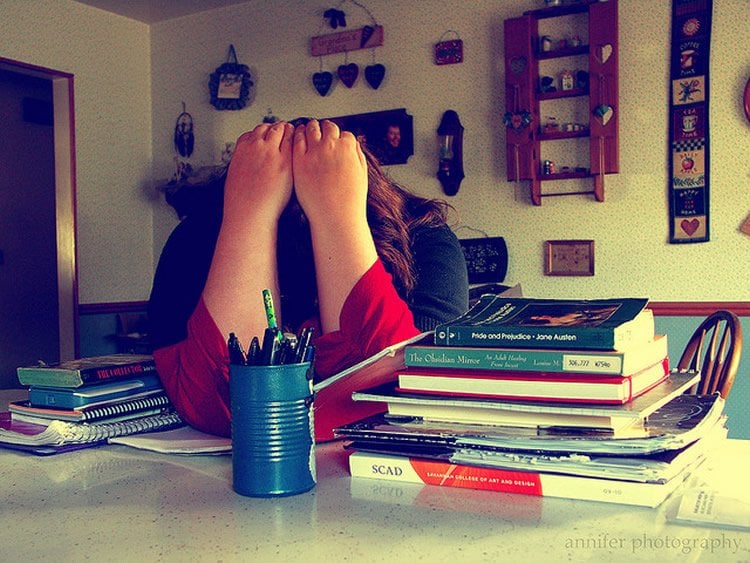 This shows a woman holding her head while resting it on a table. She is surrounded by books. The image implies the woman is stressed.
