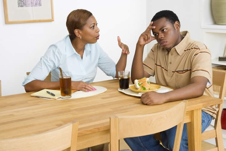 Image of a mom and son sitting at a table. The mom looks to be telling off the son, who has his head in his hands.