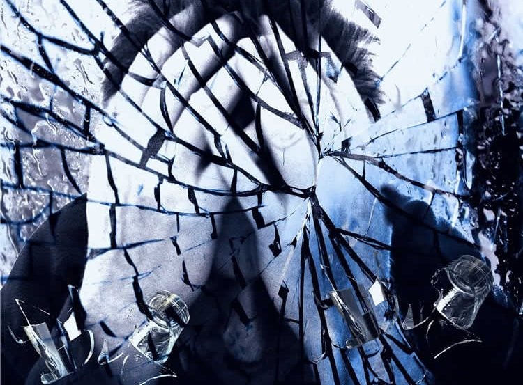 Image shows a man holding his head behind smashed glass.