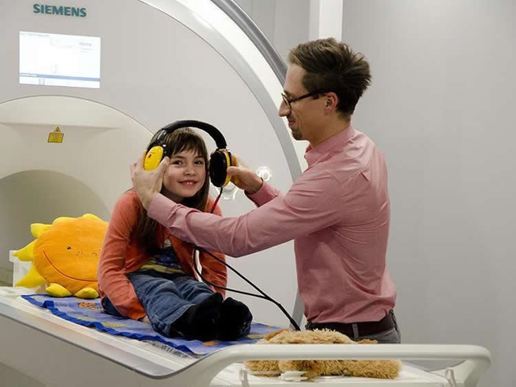 Image shows a little girl in headphones sitting on an MRI scanner bed.