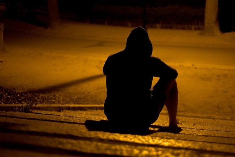 Image shows a depressed man sitting on a street in the dark.