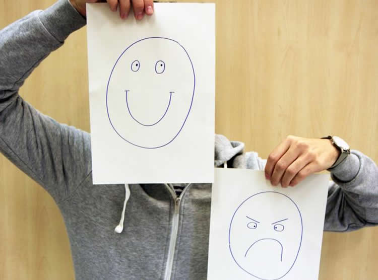 Image shows a person holding up a happy face drawing and a mad face drawing.