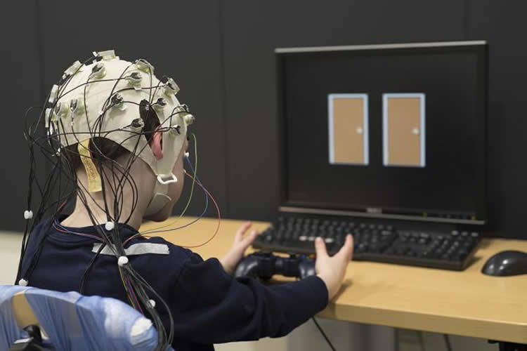 A child wearing a device that measures electrical activity in the brain chooses between doors on a computer screen. Choosing one door wins points while the other results in a loss of points. Washington University researchers have found that the brains of children with depression don't react as robustly to success in the game. Their blunted reward response is a marker of clinical depression. NeuroscienceNews image is credited to Robert Boston.