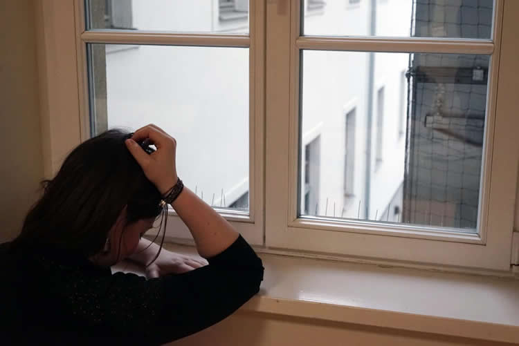 Image shows a depressed woman looking out a window.