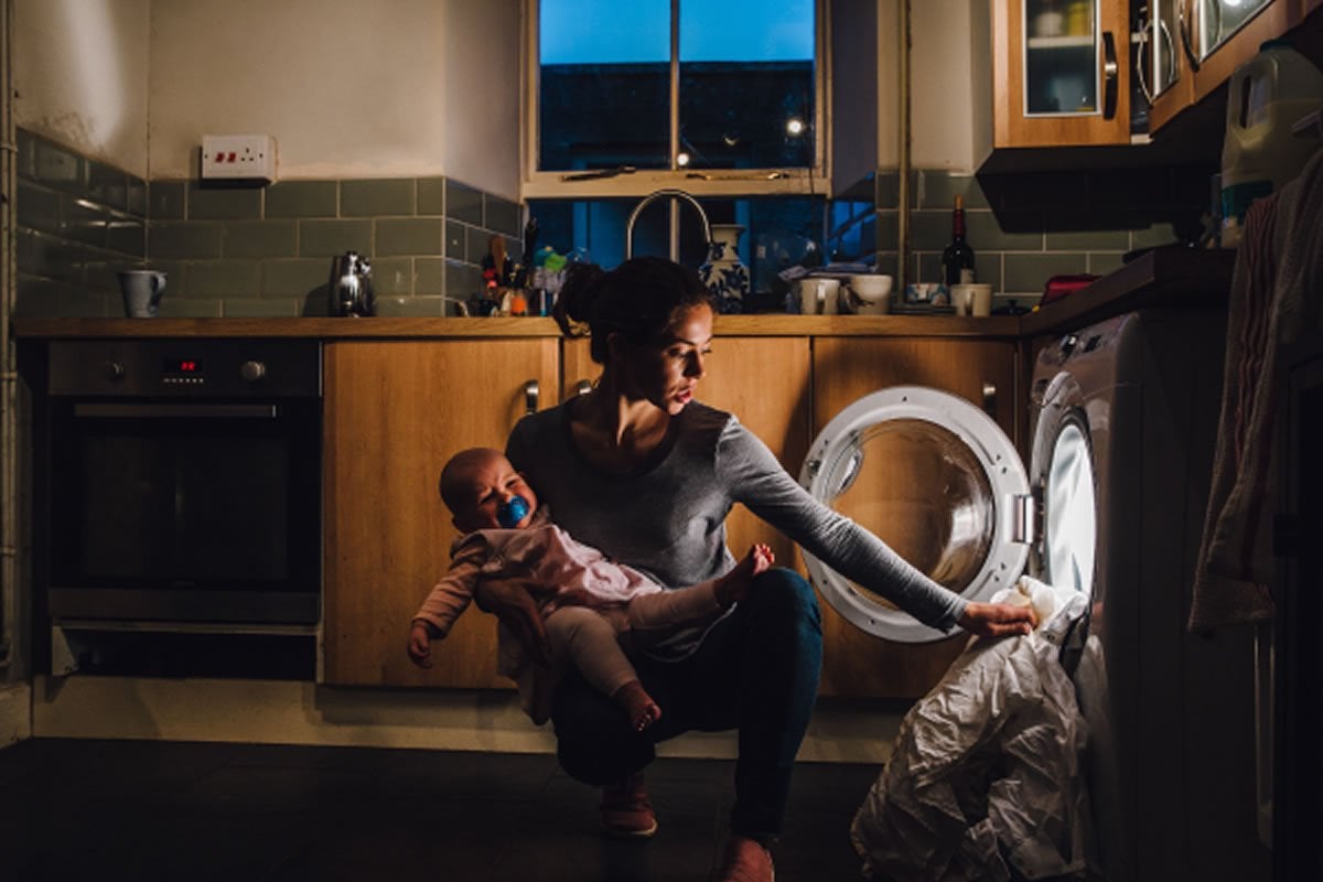 This shows an exhausted mom with a baby loading a washing machine