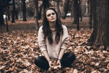 This shows a sad looking woman sitting in a forest with bare trees and leaves on the ground