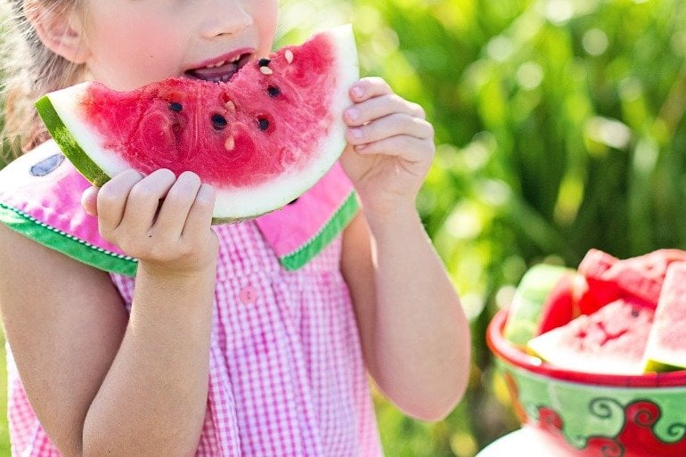 This shows a little girl lifting a piece of watermelon to her lips