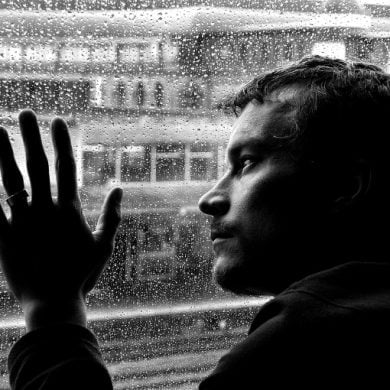 This shows a sad looking man looking out of a window at the rain