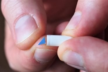 This shows a man's fingers holding a tiny blue triangle that looks as though it is made out of felt