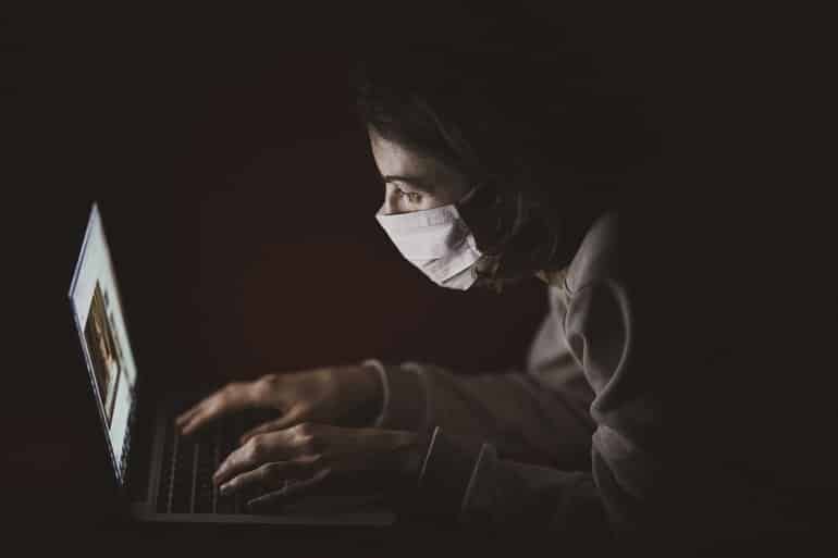 Isso mostra uma mulher com uma máscara em um quarto escuro, olhando para um laptop