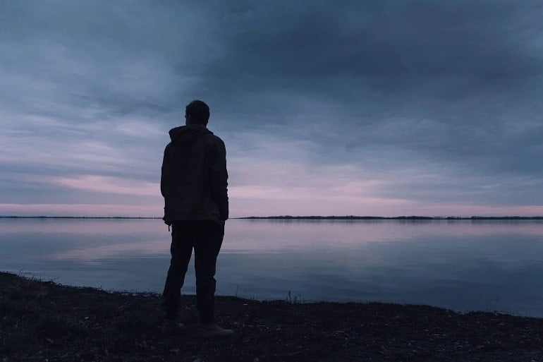 This shows a man standing alone, looking out onto a stream at night time