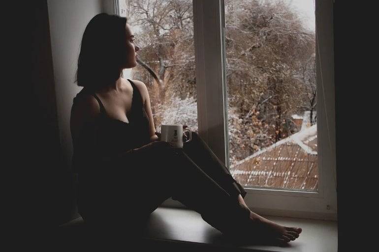 This shows a depressed looking woman looking out a window with a coffee in her hand