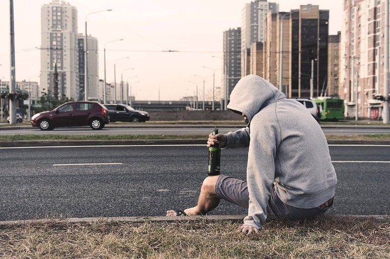 This shows a young man sitting at the side of a road with a bottle of beer