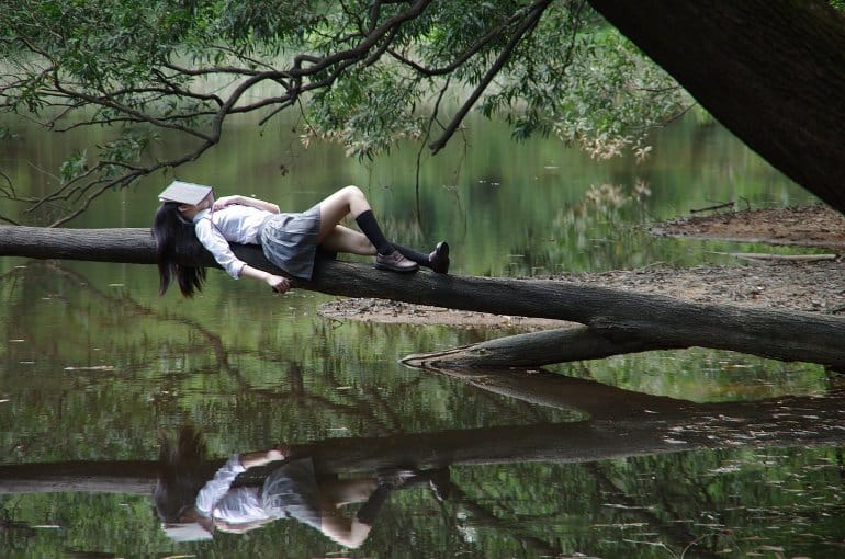 This shows a woman taking a nap on a tree limb by a lake with a book covering her face