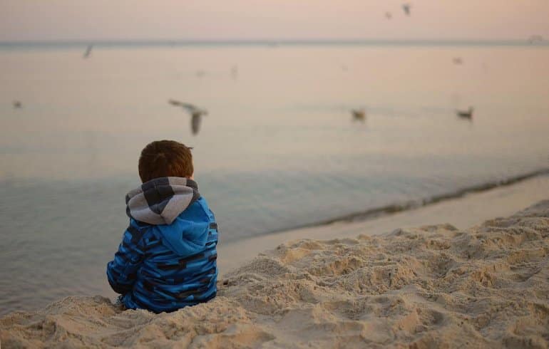 This shows a sad little boy sitting on a beach looking at the sea