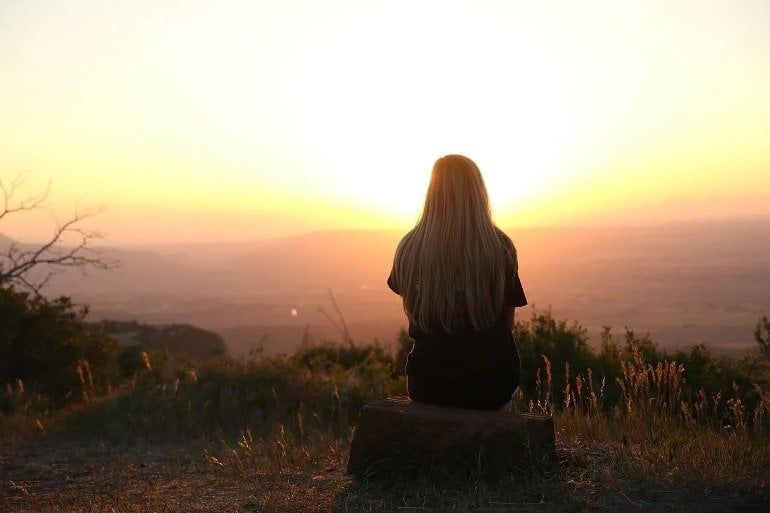 This shows a woman sitting on top of a hill, looking at a sun set