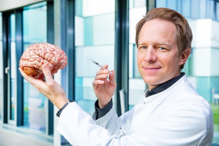 This shows the researcher holding a model of a brain and a syringe