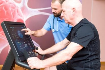 This shows an older man on an exercise bike in a hospital. A nurse is at his side. On the cycle is a monitor with a brain game.