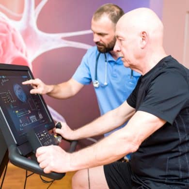 This shows an older man on an exercise bike in a hospital. A nurse is at his side. On the cycle is a monitor with a brain game.