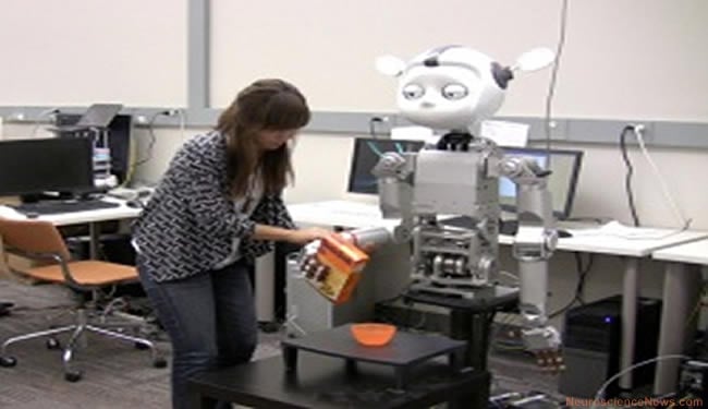 A woman is holding the hand of a robot as it appears to pour something from a box into a bowl on a table. The robot's eyes appear to be half open.